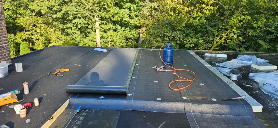 Pose d'étanchéité en cours sur toit plat : rouleau de membrane bitumineuse à moitié déroulé au centre, bouteille de gaz propane bleue et chalumeau posés sur la lé.